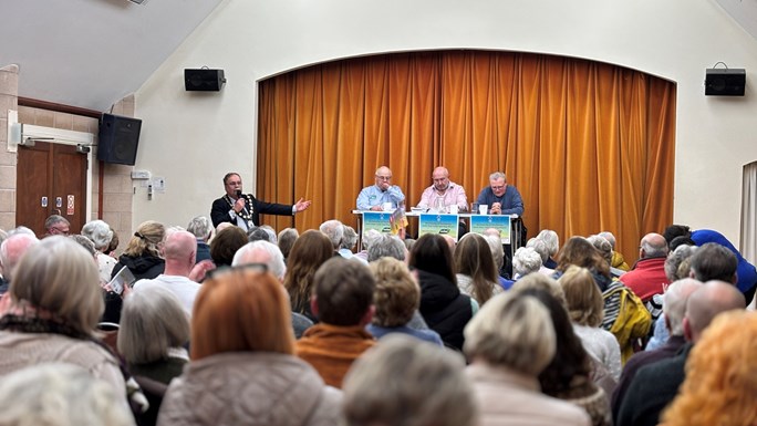 view from back of a room with a stage at the front and 3 males sat on the stage